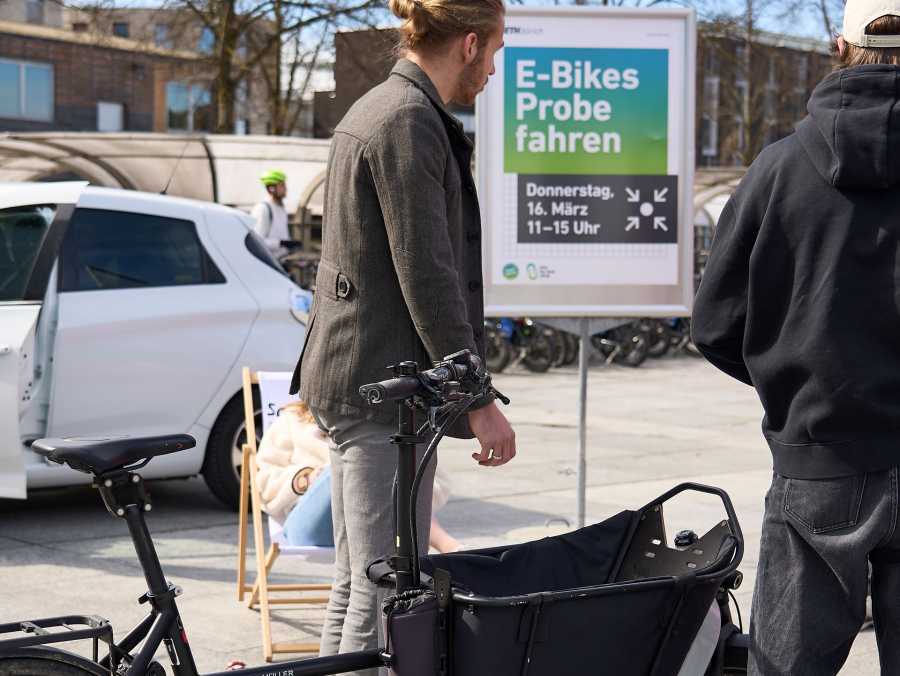 Vergr?sserte Ansicht: Menschen mit Velo auf dem ETH Gel?nde und Plakat im Hindergrung zum Showcase Bikesharing