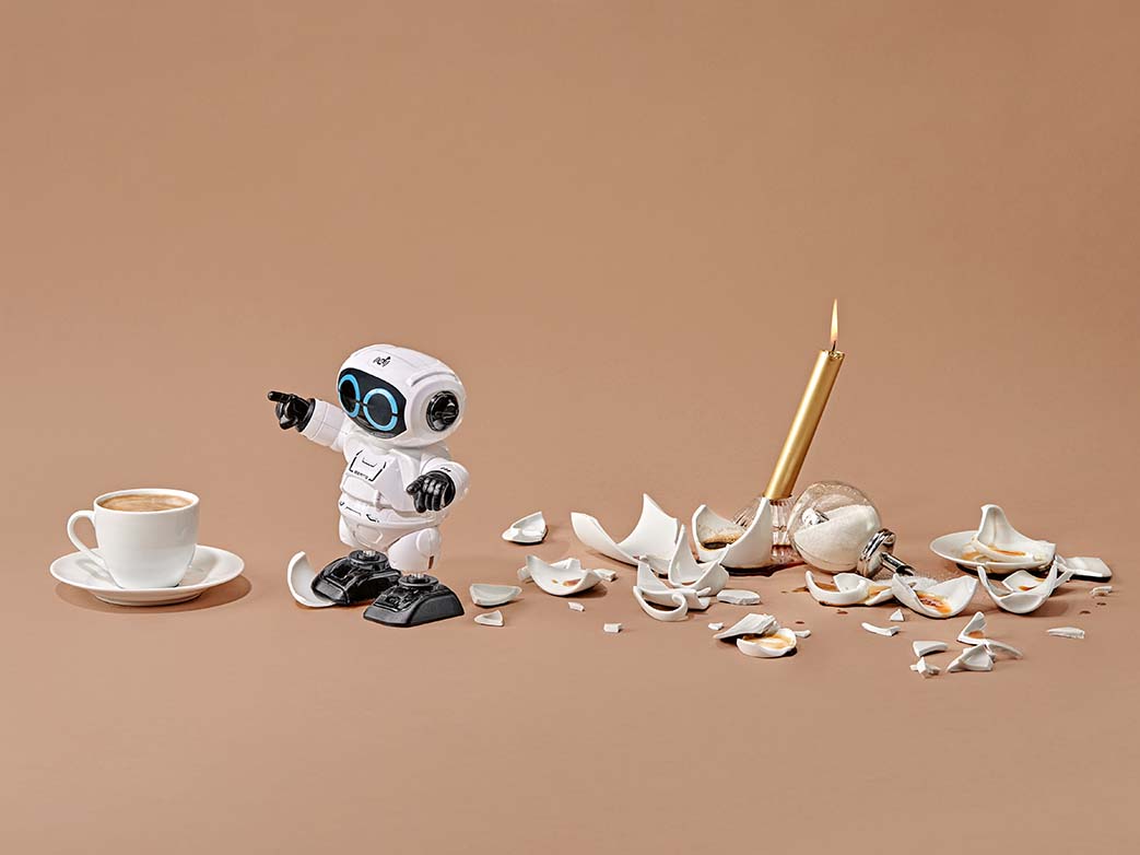Ein Mini-Roboter neben einer kaputten Kaffeekanne, einer Kaffeetasse, einer Kerze und einem Zuckerspender