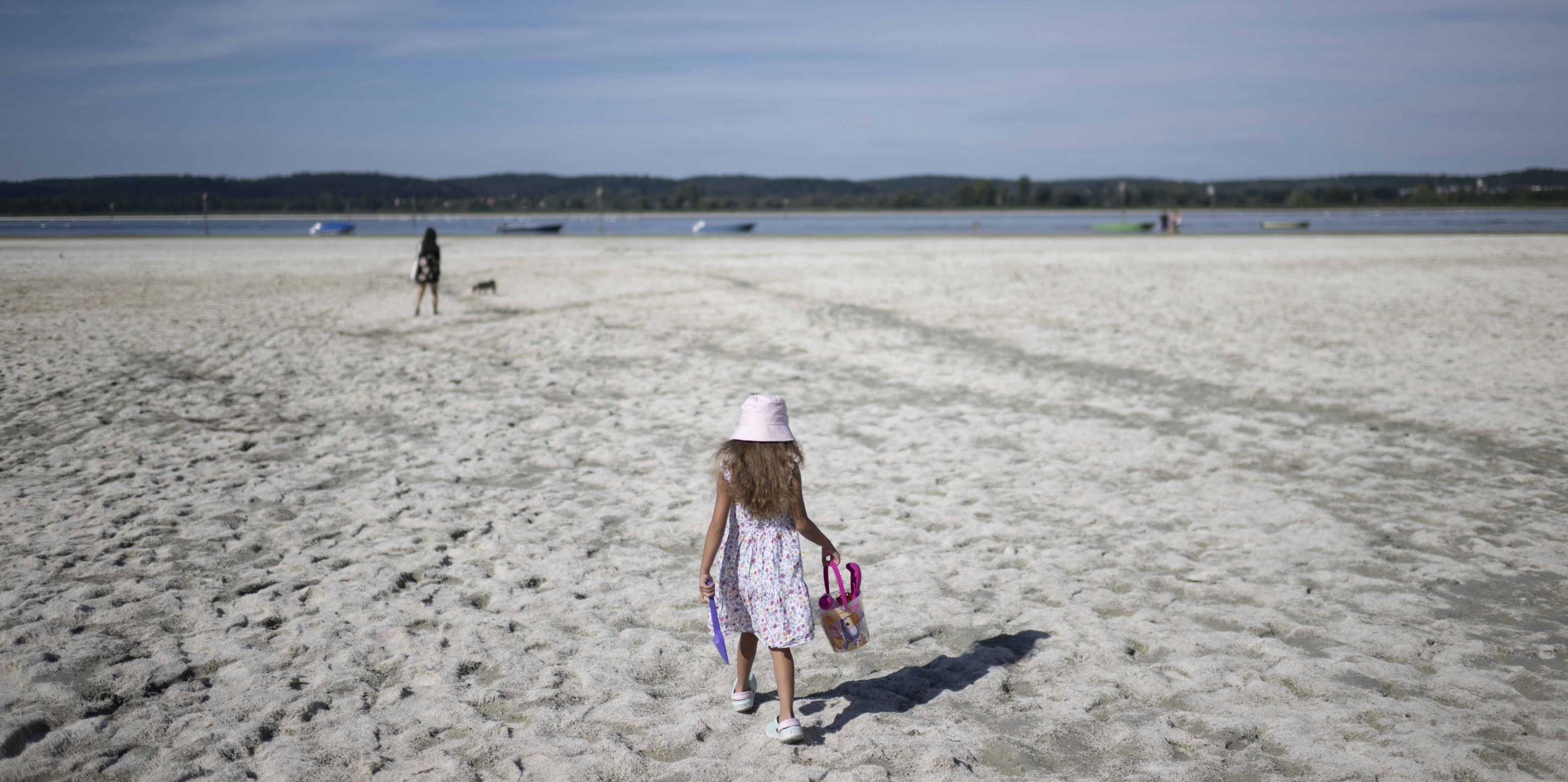 Ein kleines Mädchen geht auf Sand, in der Hand hat sie ein Kesselchen und eine Schaufel. Im Hintergrund ist eine Frau mit einem Hund und das Meer zu erkennen.