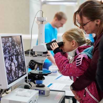 Ein kleines Mädchen wird von ihrer Mutter gehalten, während es konzentriert durch ein Mikroskop blickt.