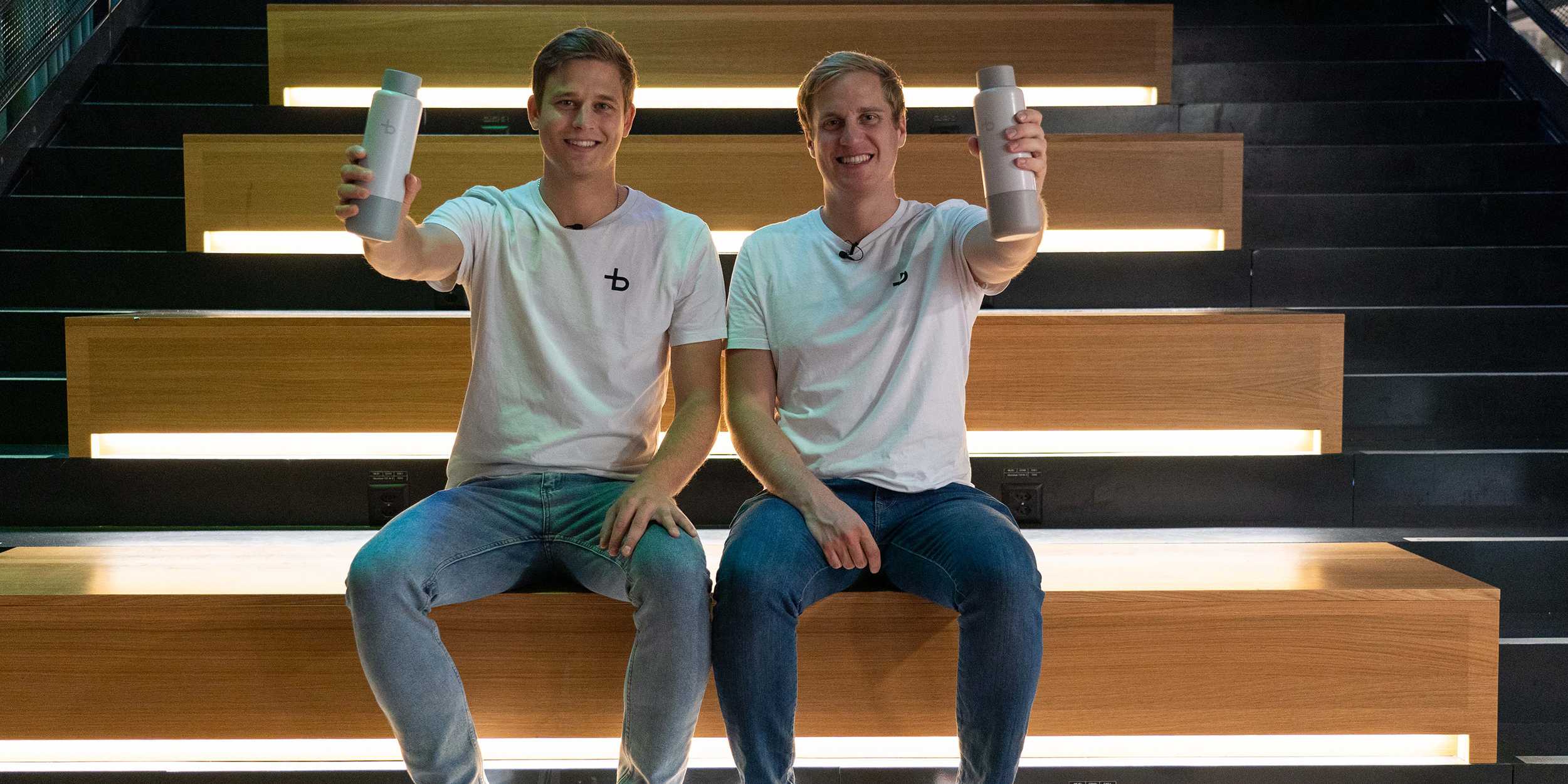 Christian Käser und Linus Lingg sitzen auf einer Treppe und halten ihre Bottleplus Flasche in die Höhe.