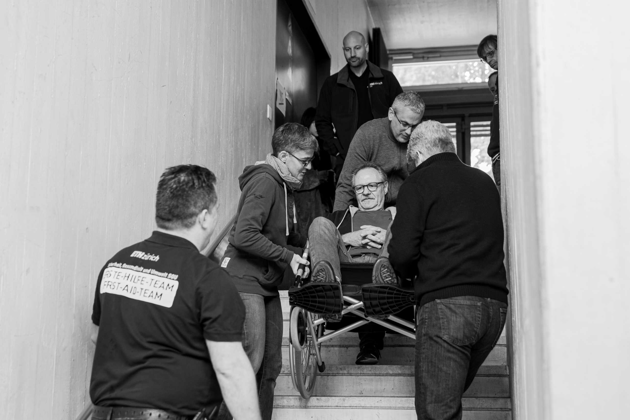 Eine Gruppe von Personen evakuiert einen Mann im Rollstuhl über das Treppenhaus. 