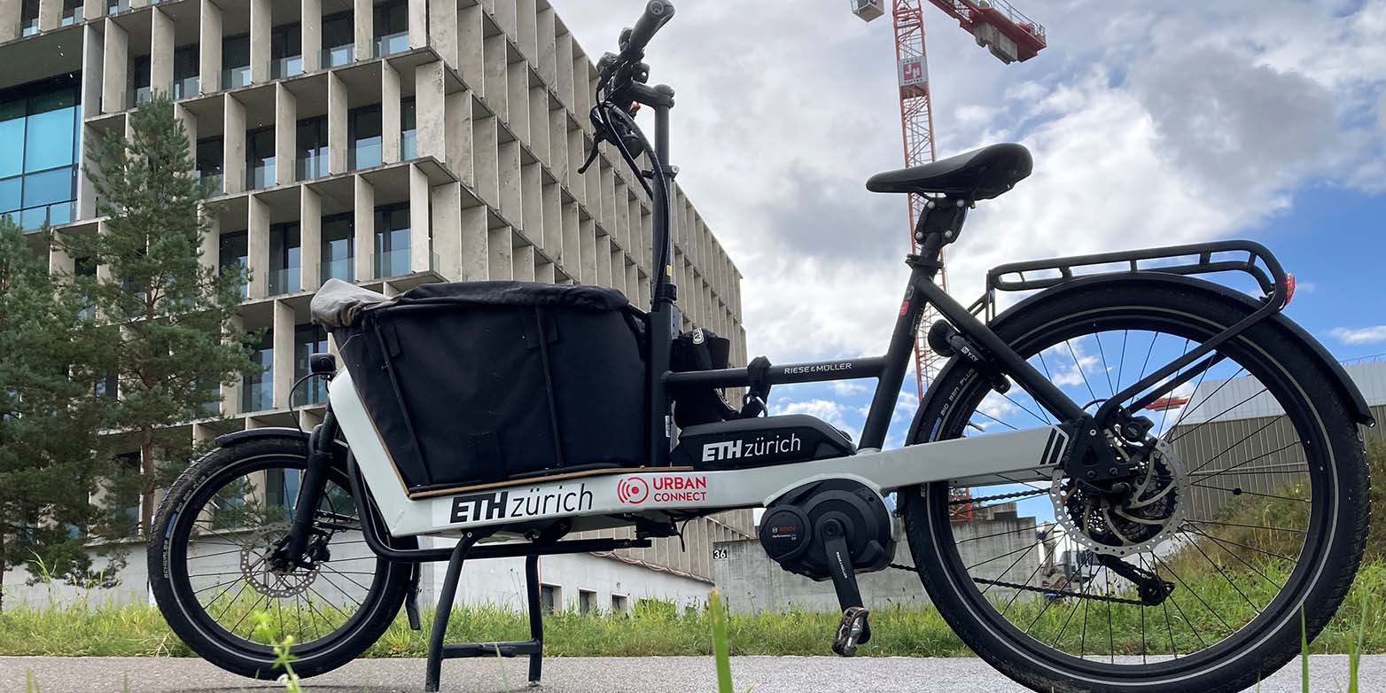 Das E-Cargo-Bike vor einer Wiese, mit Gebäude und Kran