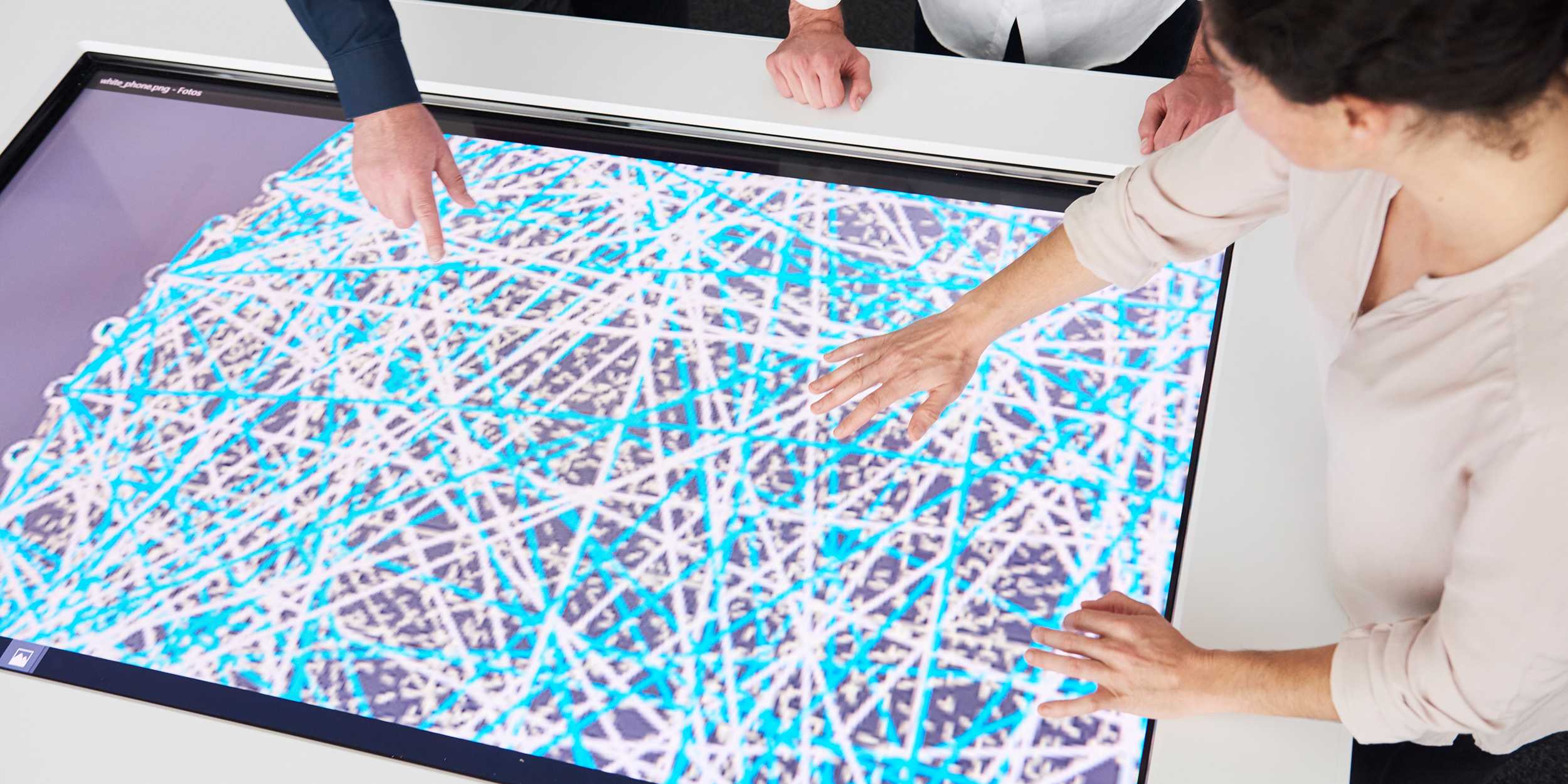 Zwei Personen zeigen mit ihren Händen auf einen Bildschirm auf dem ein Netzwerk aus weissen und blauen Linien zu erkennen is.
